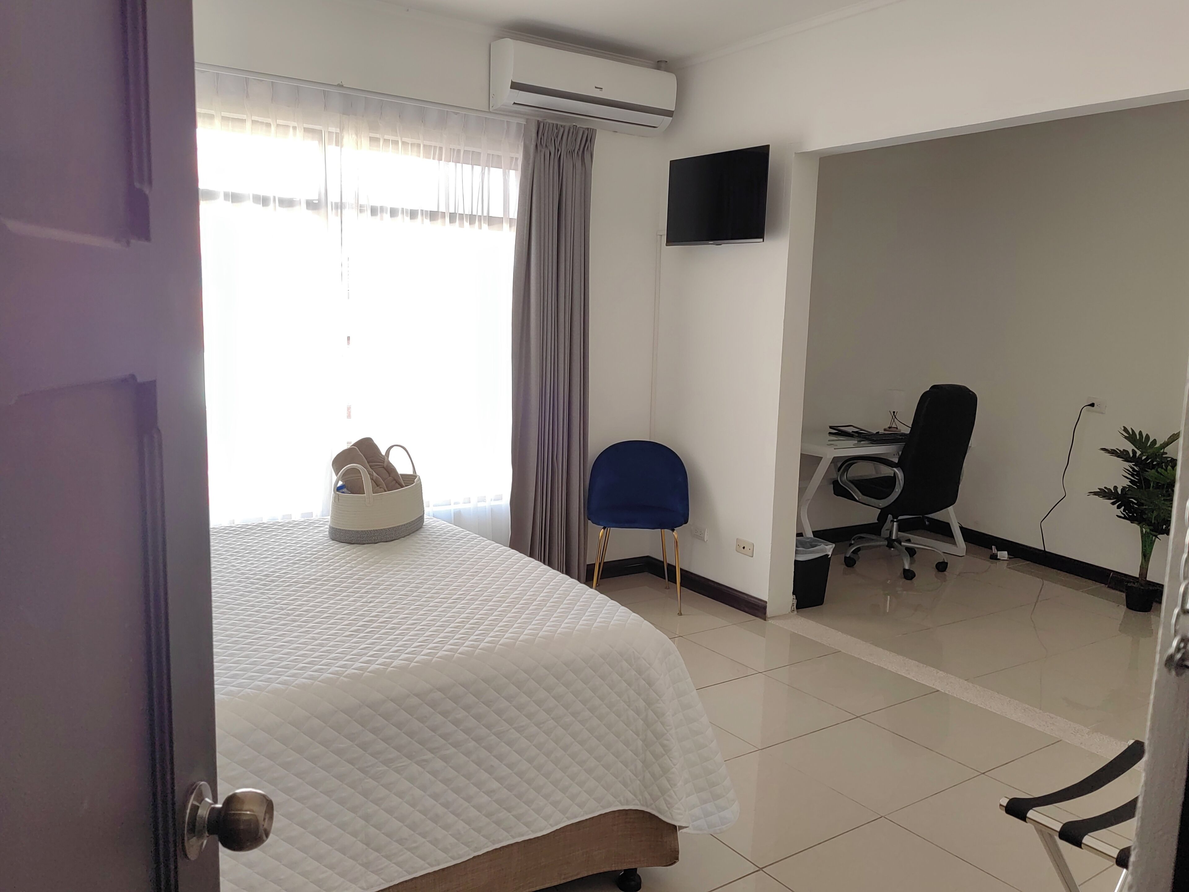 A hotel room with a bed, a chair, a desk with a computer, and a TV on the wall.