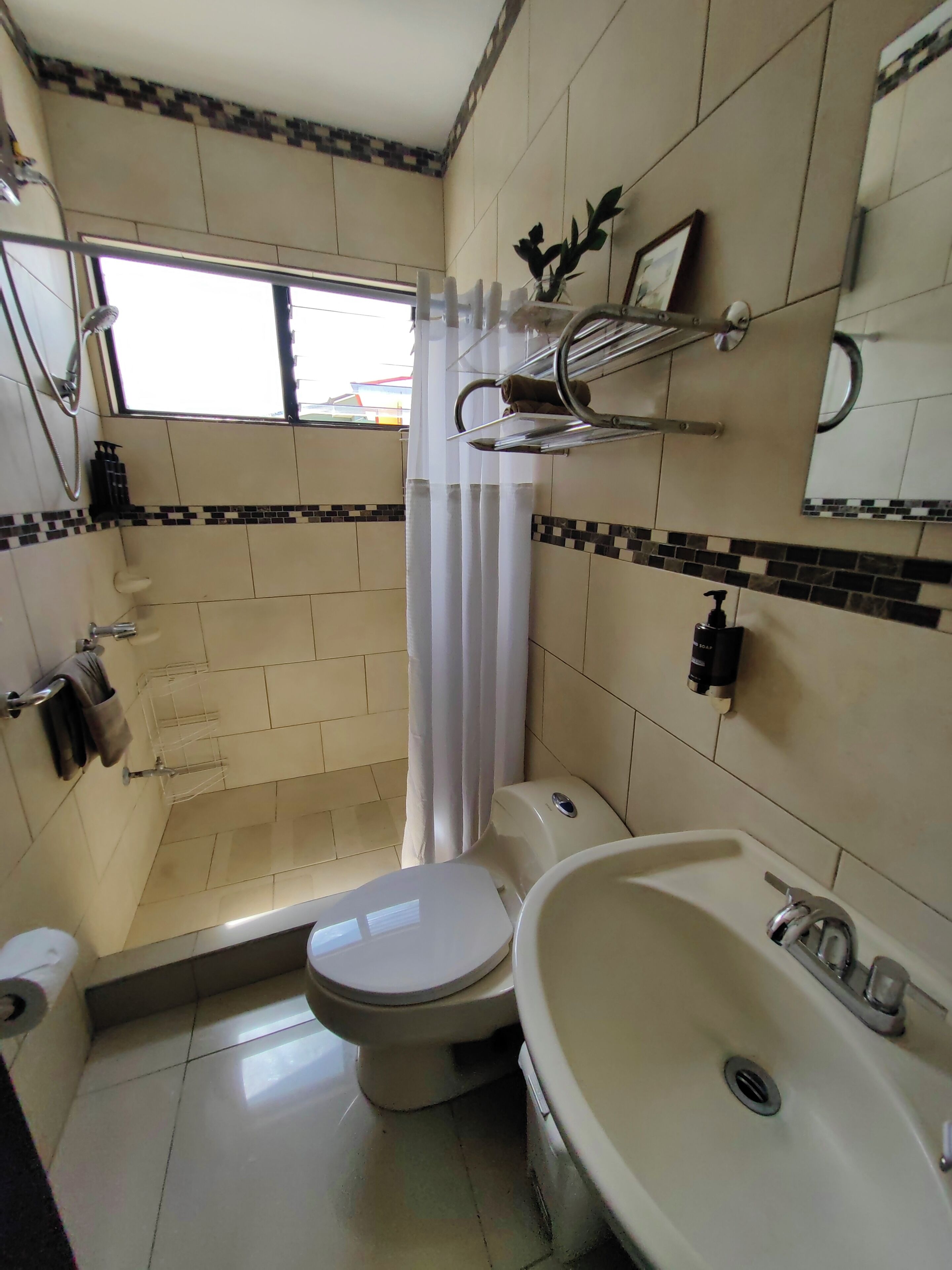 A small bathroom with a toilet, sink, and shower. There is a window providing natural light and a towel rack.