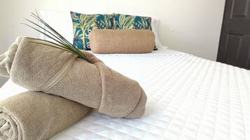 Een net opgemaakt bed met een wit gewatteerd sprei, twee beige handdoeken, een kussen met blauw- en groen patroon, en een beige kussen.