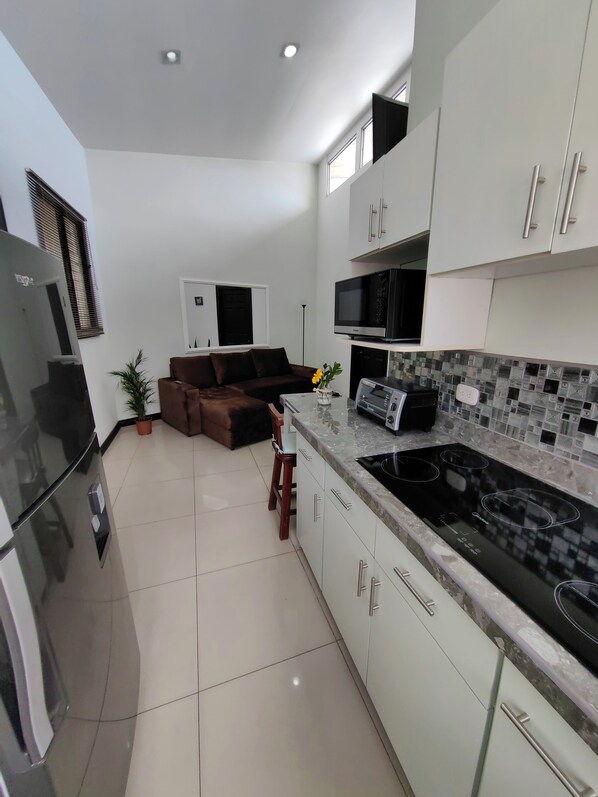 A modern kitchen with white cabinets, a black stove, and a microwave. There is a brown sofa and a potted plant in the living area. - Hotel Boutique CasAguacate (La Asuncion)