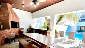 Un espacio exterior para comer con horno de ladrillo, bancos de madera, ventilador de techo y vista a una piscina y cielo azul.