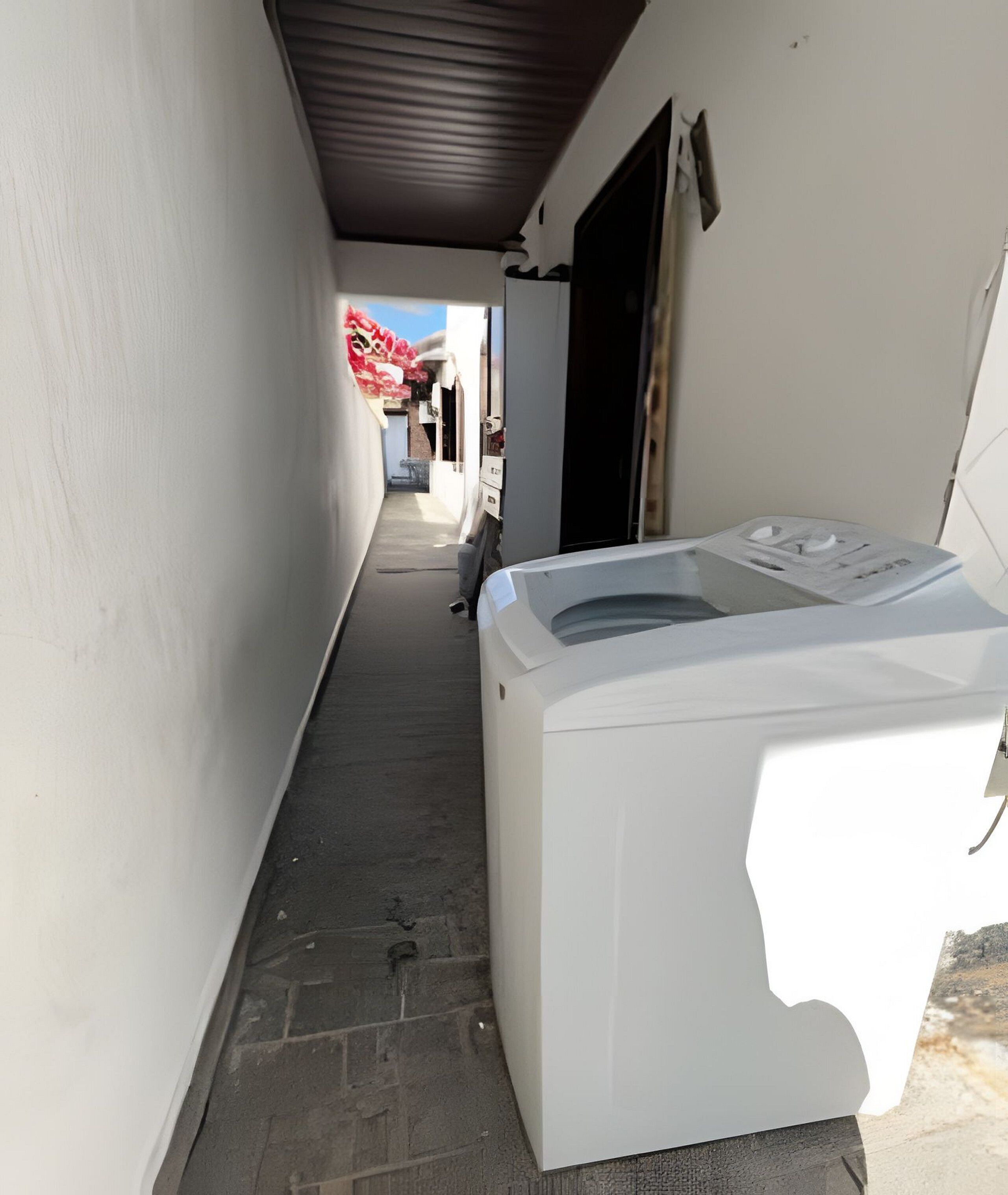 A narrow outdoor corridor with a white washing machine on the right, a door on the left, and a red awning in the distance.