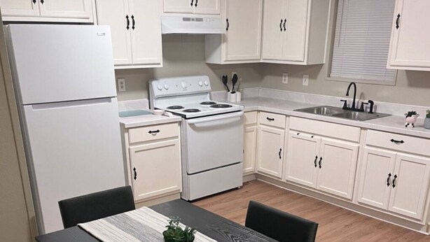 Une cuisine compacte avec des meubles blancs, un réfrigérateur blanc, une cuisinière et un évier. On y trouve une table à manger avec des chaises et une plante posée sur la table.