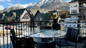 A patio with a round glass table and chairs, a grill, and a view of snow-capped mountains.