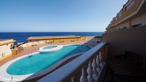 A pool area with a view of the ocean, lounge chairs, and a building with balconies.