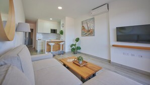 A modern living room with a sofa, a wooden coffee table with a plant, a TV mounted on the wall, and a kitchen in the background.