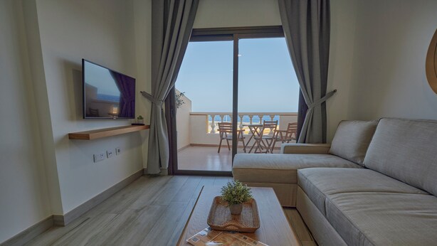 A modern living room with a sofa, a coffee table with a newspaper, a TV on the wall, and a balcony with a view of the sea.
