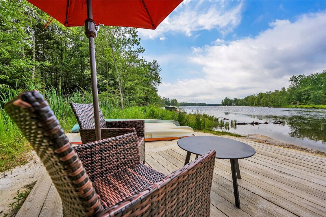 Een houten terras met rieten stoelen en een tafel, met uitzicht op een rustig meer met boten, omgeven door weelderige begroeiing.
