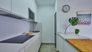 A narrow kitchen with white cabinets, a black countertop, a wooden cutting board, a sink, a microwave, a clock, and a painting on the wall.