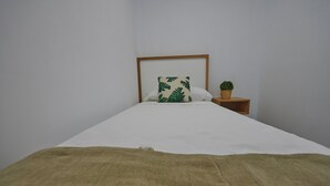 A neatly made bed with a white sheet and a green leaf-patterned pillow, a small wooden nightstand with a potted plant, and a plain white wall.