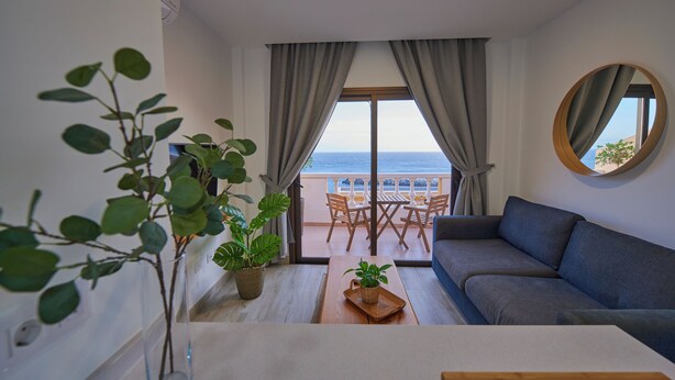 Een moderne woonkamer met een bank, een eettafel en uitzicht op zee door grote ramen.