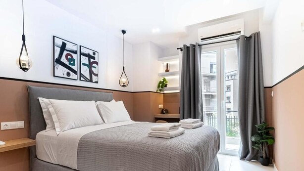 A modern bedroom with a bed, bedside tables, a nightstand, a potted plant, and a view of the outside balcony. - Mar25 #31 by Smart Cozy Suites (Athina)