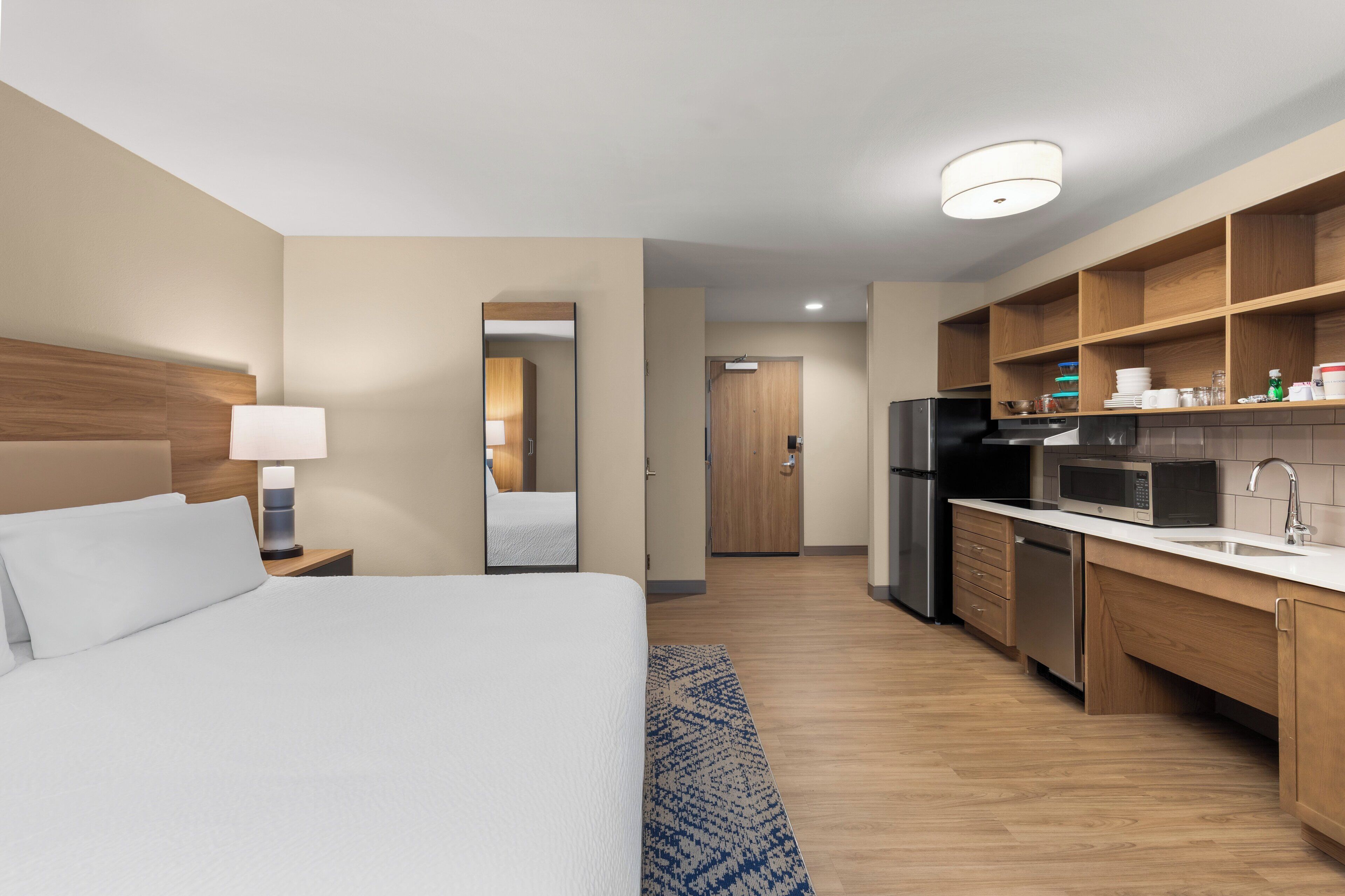 Habitación de hotel con cama, mesita de noche, lámpara y vista al área de la cocina.