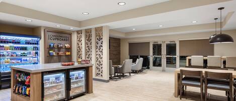 Un moderno vestíbulo de hotel con un expositor refrigerado, una zona de estar con mesa y sillas, y una pared decorativa con motivos geométricos.
