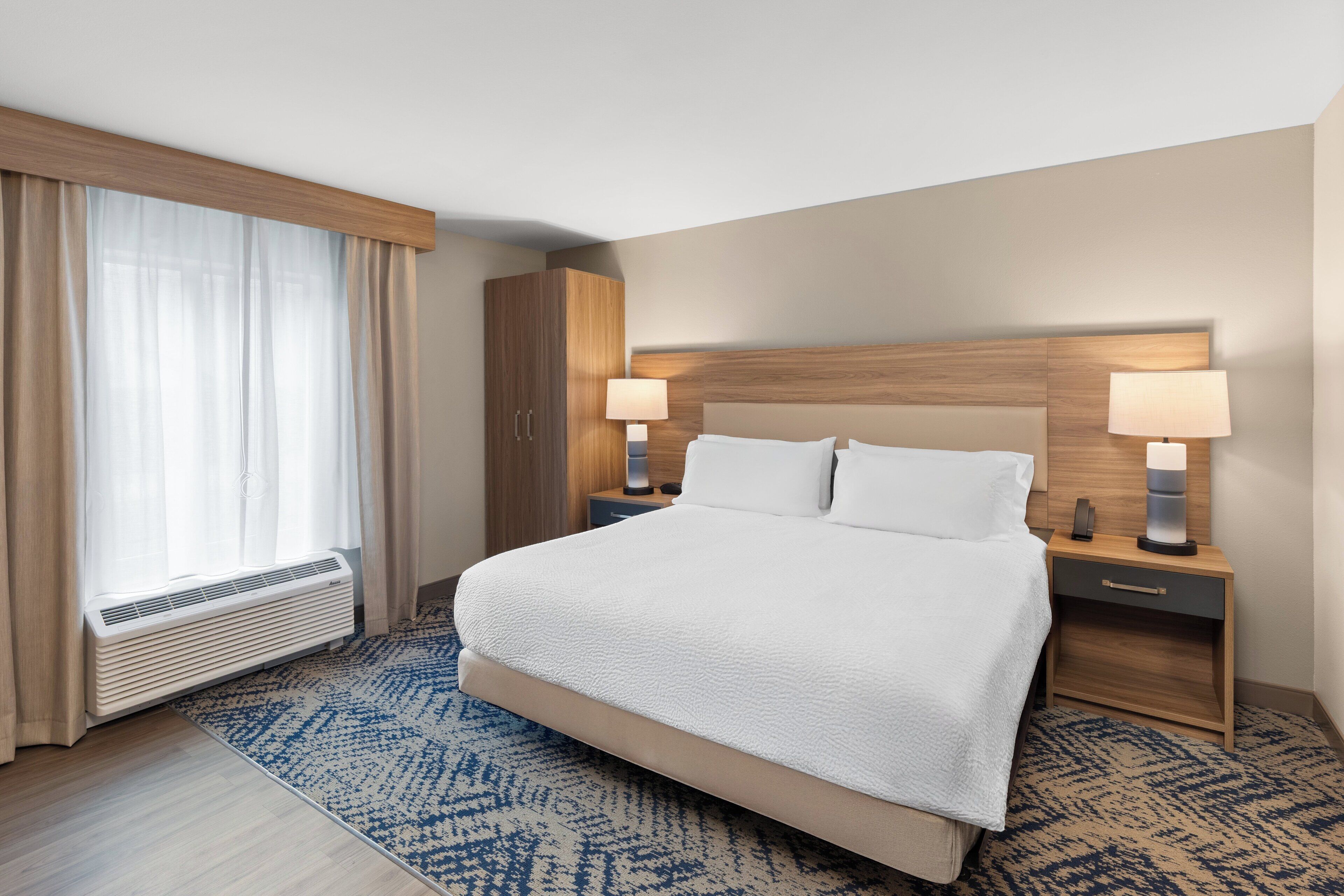 Een hotelkamer met een groot bed, twee nachtkastjes met lampen, een houten hoofdbord, een raam met dunne gordijnen en een vloerverwarming.
