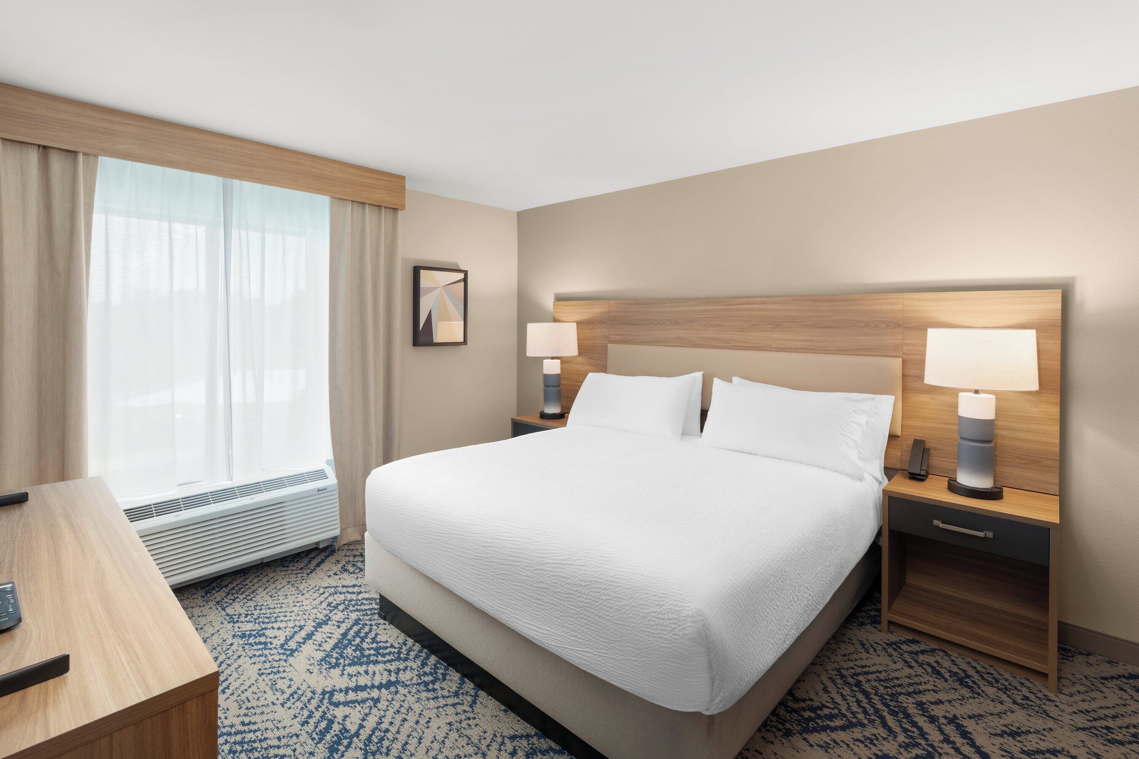 A hotel room with a large bed, two bedside lamps, a desk with a chair, and a large window with curtains.