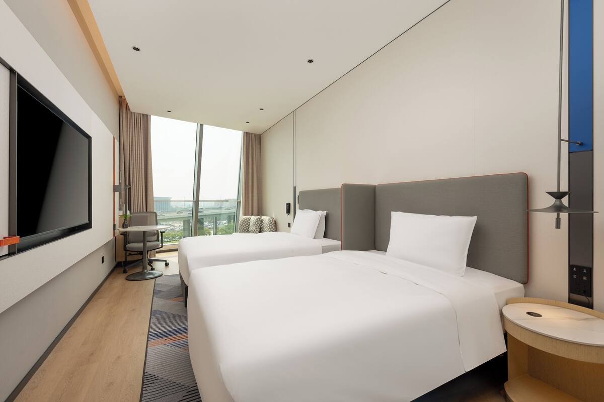 Ein Hotelzimmer mit zwei Betten, einem Fernseher, einem Schreibtisch und Blick auf die Stadt.