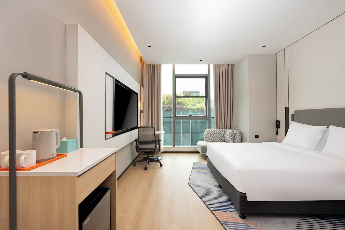 Ein modernes Hotelzimmer mit einem Bett, einem Schreibtisch mit Flachbildfernseher, einem Sessel und einem Fenster mit Blick ins Freie.