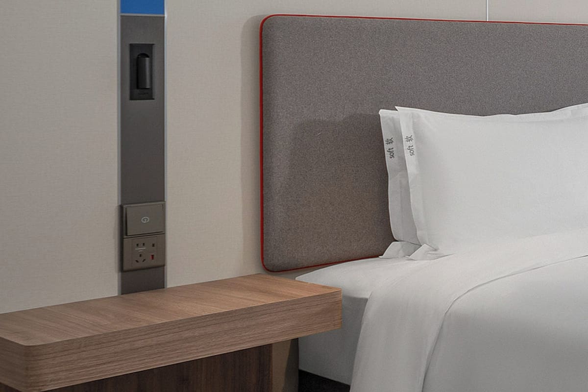 Une chambre d’hôtel moderne avec une table de chevet en bois, une tête de lit grise et un lit blanc.