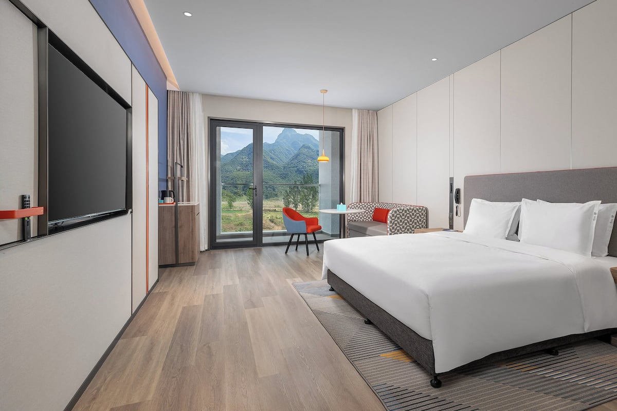 Une chambre d’hôtel moderne avec un grand lit, une petite table avec des chaises, une télévision et une vue sur les montagnes, visible à travers la porte-fenêtre coulissante.