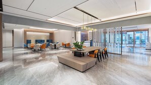 Un hall d’hôtel moderne doté d’un espace salon central, d’un comptoir d’accueil et d’une grande porte vitrée donnant sur l’extérieur.