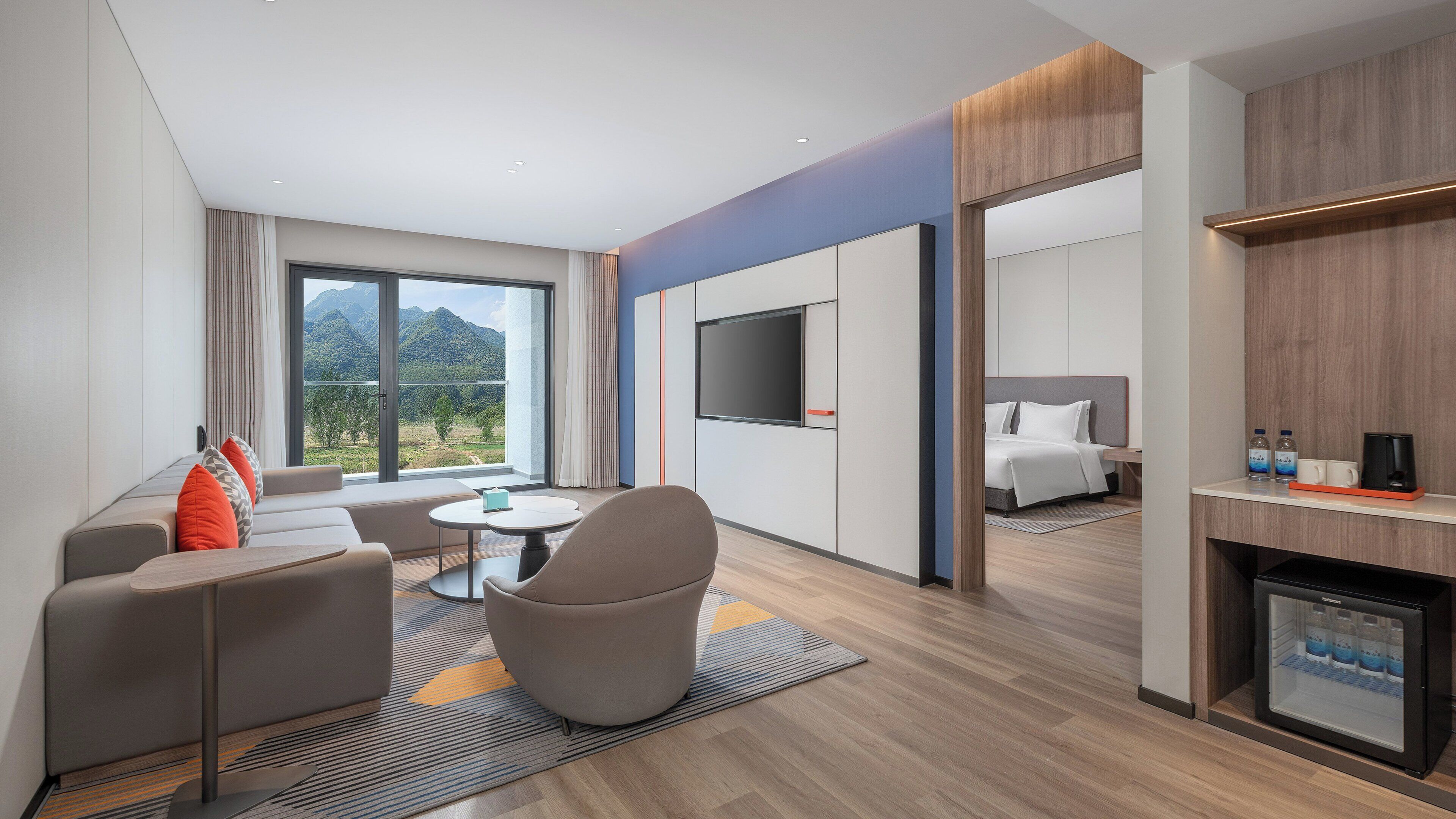 A modern hotel room with a large window, a flat-screen TV, a sofa, a coffee table, and a bed.