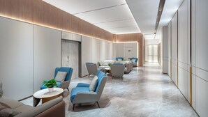 Un hall d’hôtel moderne avec un long couloir, des espaces de détente dotés de fauteuils bleus et gris, et un sol en marbre.