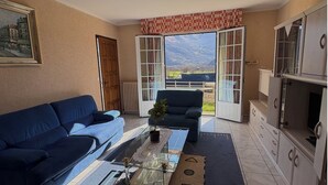 A living room with a sofa, armchair, and coffee table, a kitchen with cabinets, and a large sliding glass door leading to a balcony with a mountain view.