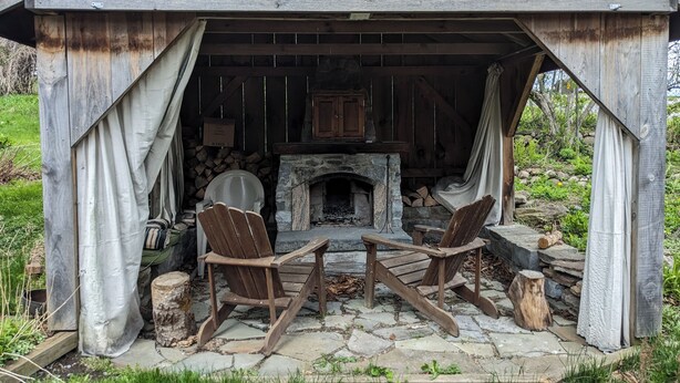 A rustic outdoor seating area with a stone fireplace, wooden chairs, and a covered porch.