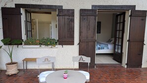 Une terrasse extérieure avec une table ronde et des chaises, un pot de fleurs et une fenêtre avec des volets en bois.