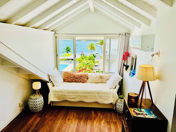 Ein Schlafzimmer mit Bett, Fenster mit Blick auf Pool und Palmen, Lampe und kleiner Tisch mit Buch.