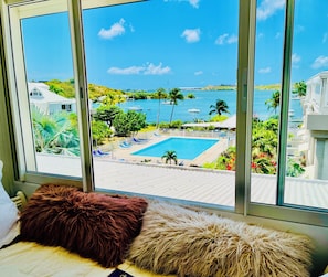 Ein Zimmer mit großem Fenster mit Blick auf einen Swimmingpool und ein Gewässer, mit Aussicht auf ein weißes Gebäude und Grünflächen.