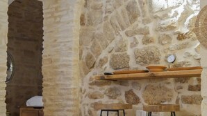 A rustic stone interior with a wooden table, two metal stools, and a shelf with pottery.