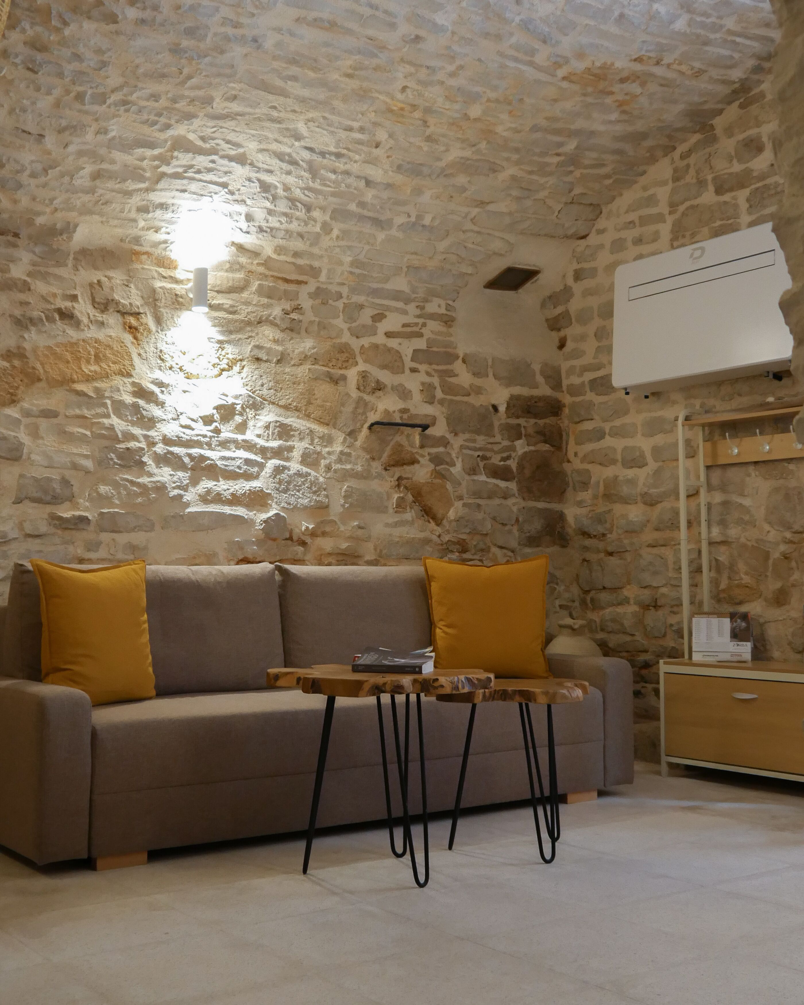A room with a stone wall, a sofa with yellow cushions, a small wooden table, and a nightstand with a lamp.