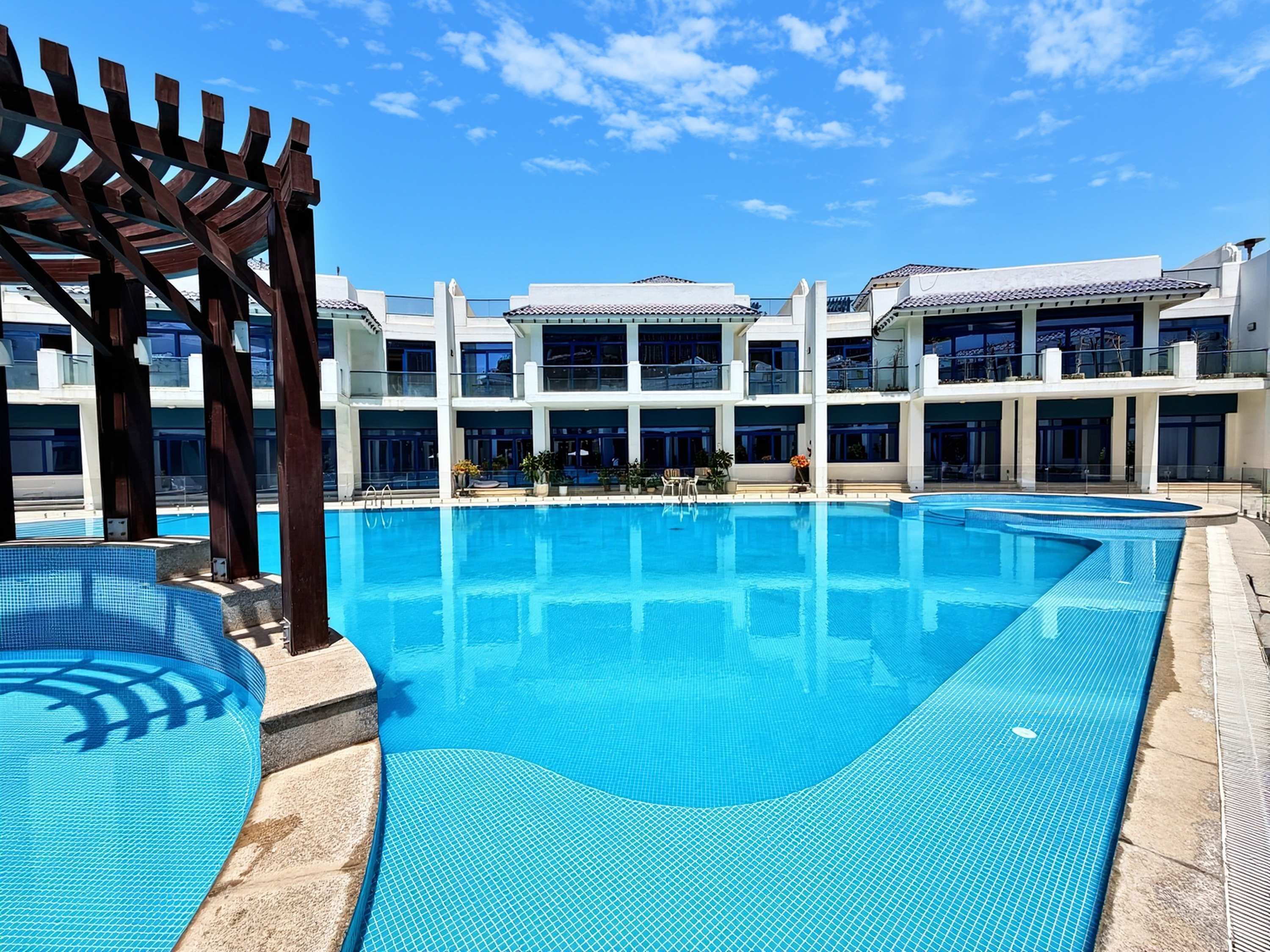 A swimming pool with a wooden pergola, a building with multiple balconies, and a clear blue sky.