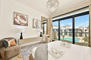 A hotel room with a bed, a sofa, a large window with a view of a swimming pool and buildings, and a chandelier.