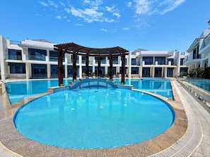 A large outdoor swimming pool surrounded by modern buildings with balconies. - 5 Bed Villa,2 Private Beaches, Pool by Estatesmen (Dubai)