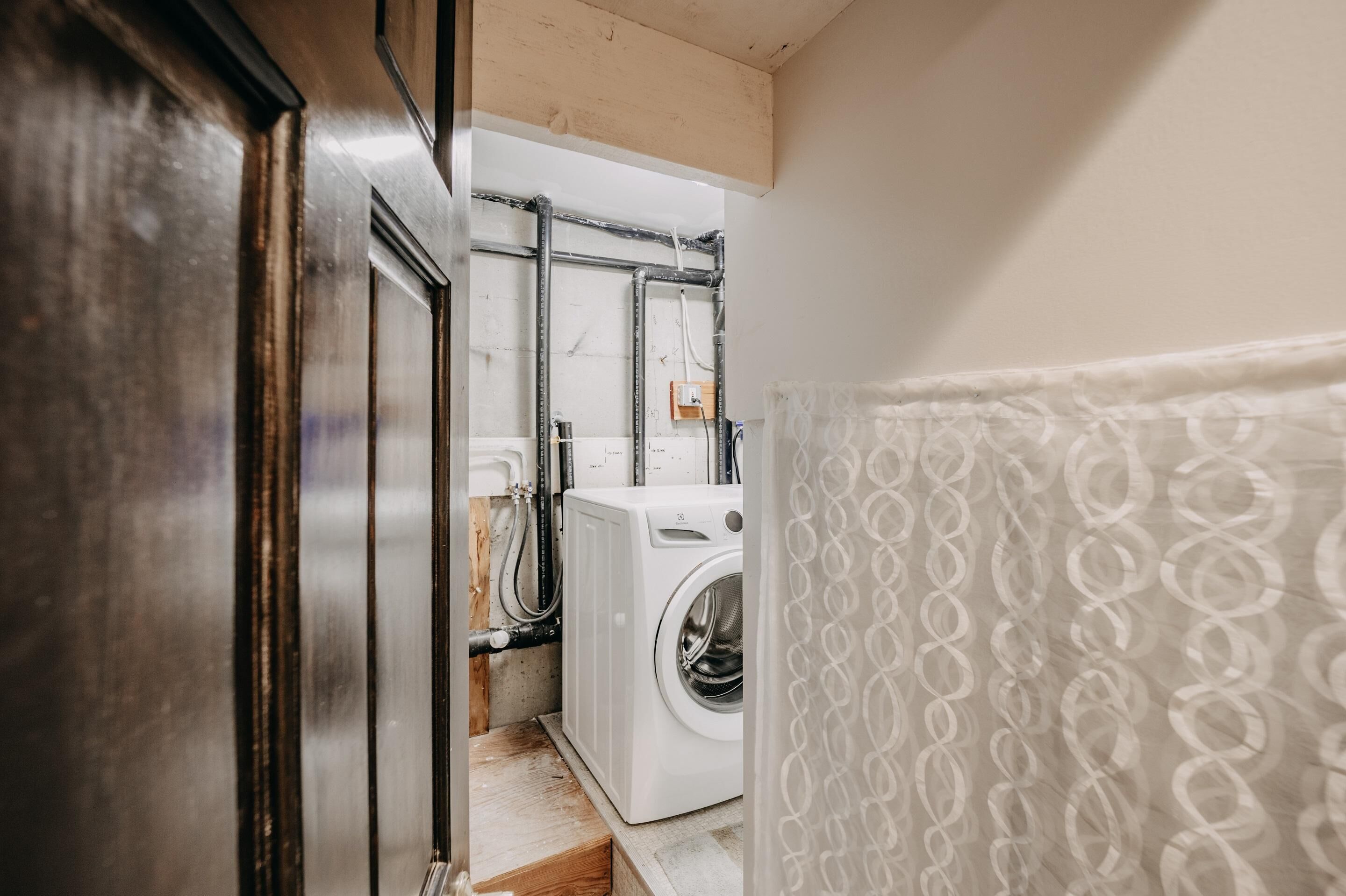 A laundry room with a washing machine, pipes, and a wooden floor.