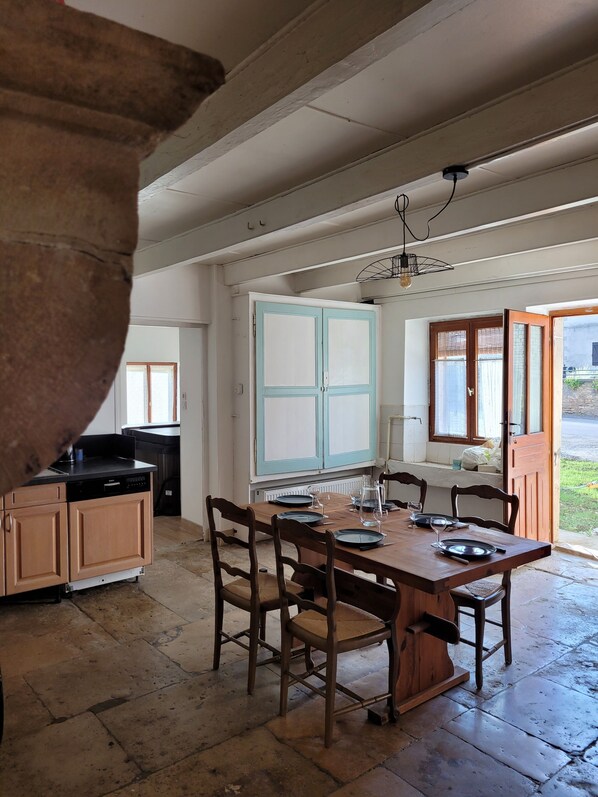 A dining area with a wooden table and chairs, a kitchen in the background, and a window with a view of the outside.