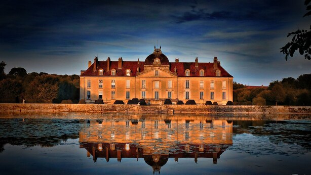 Ein großes, symmetrisches Gebäude mit einer zentralen Kuppel und zahlreichen Fenstern, das sich in ruhigem Wasser spiegelt.