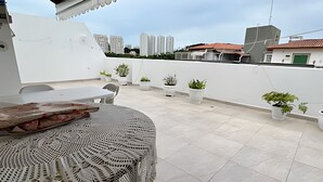 A rooftop terrace with a table and chairs, potted plants, and a view of city buildings. - Cobertura Duplex no Guarujá SP - 4 (Guaruja)