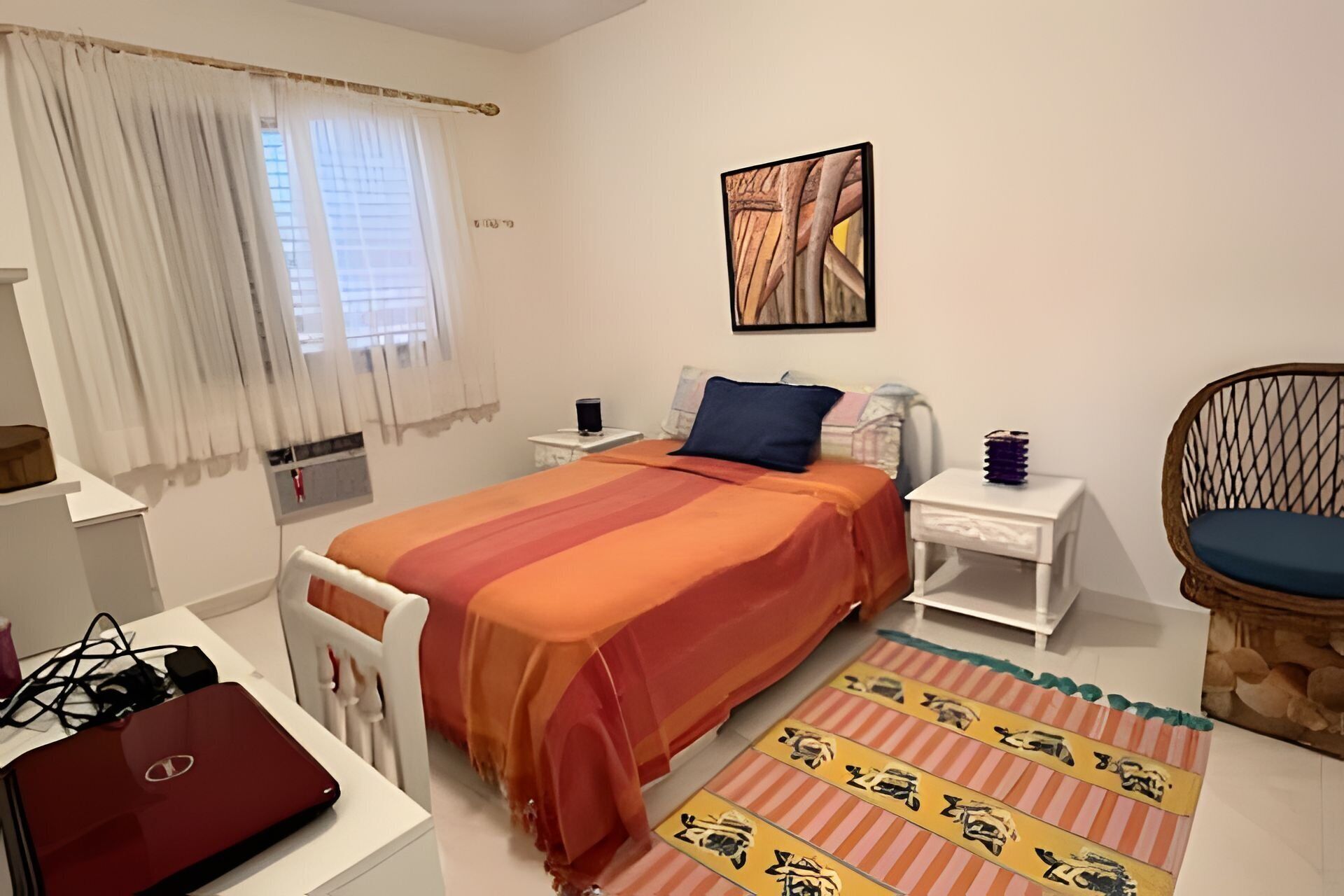 A bedroom with a bed, a desk with a laptop, a chair, a bedside table with a lamp, and a window with curtains.