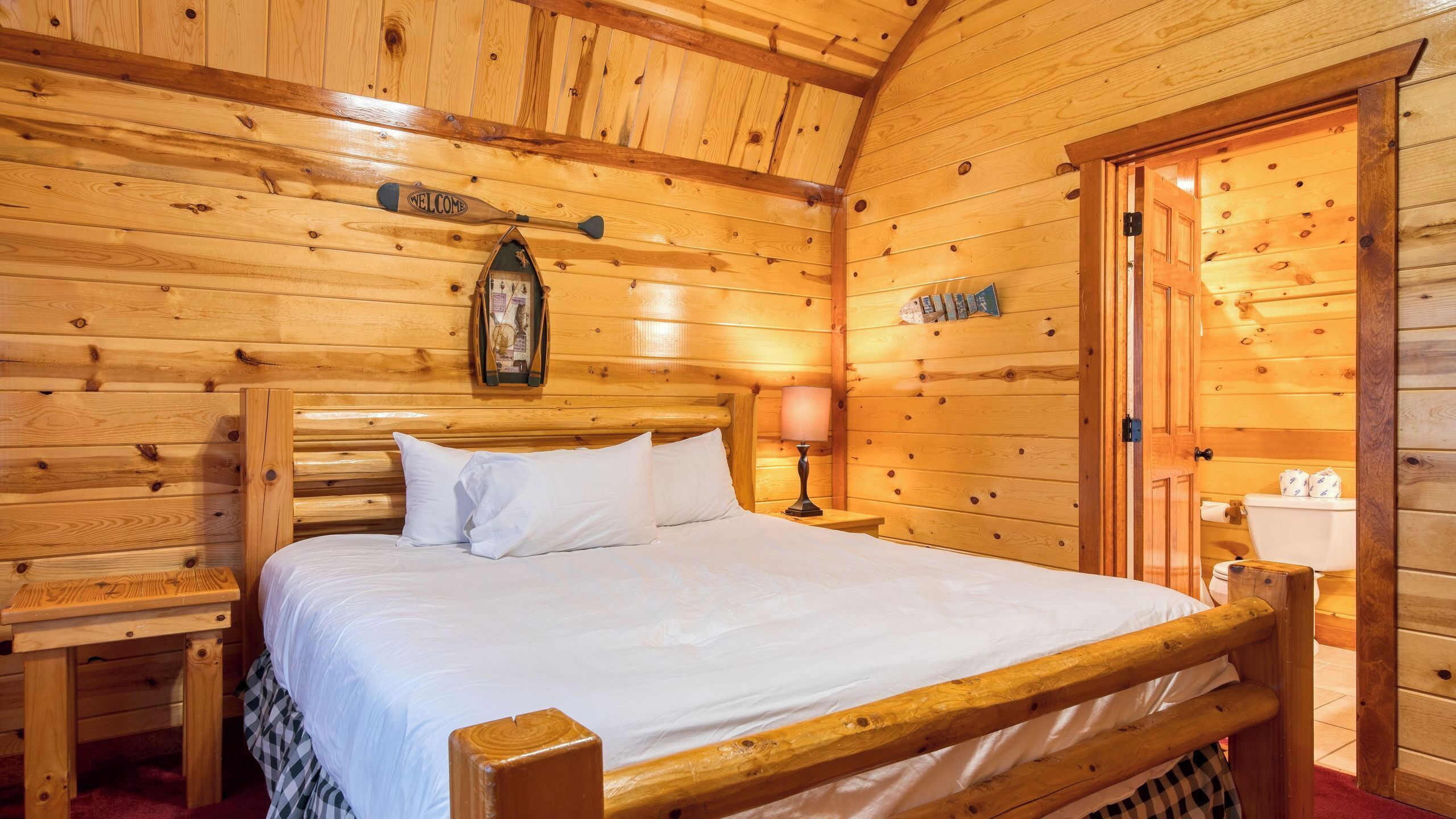 A wooden cabin-style room with a bed, a nightstand, a lamp, and a 'Welcome' sign.