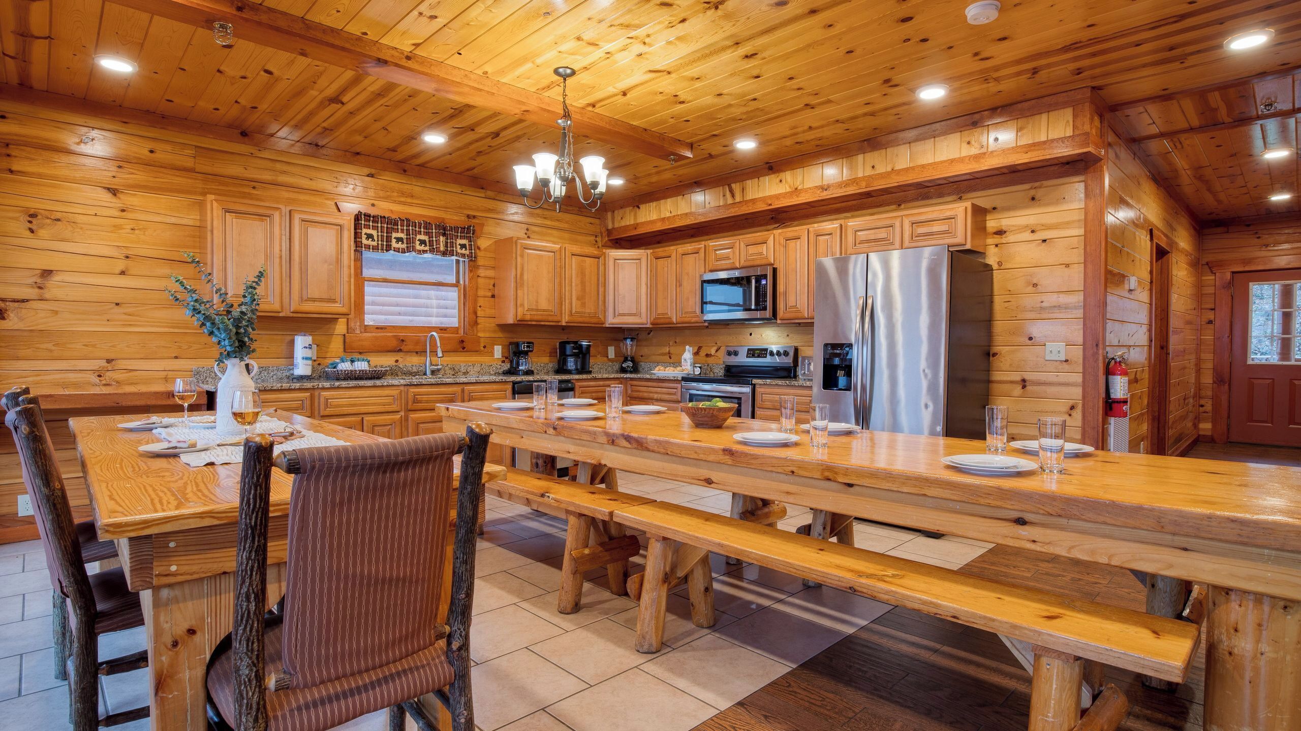 A wooden cabin-style kitchen with a long dining table, wooden chairs, a built-in microwave, and a stainless steel refrigerator.