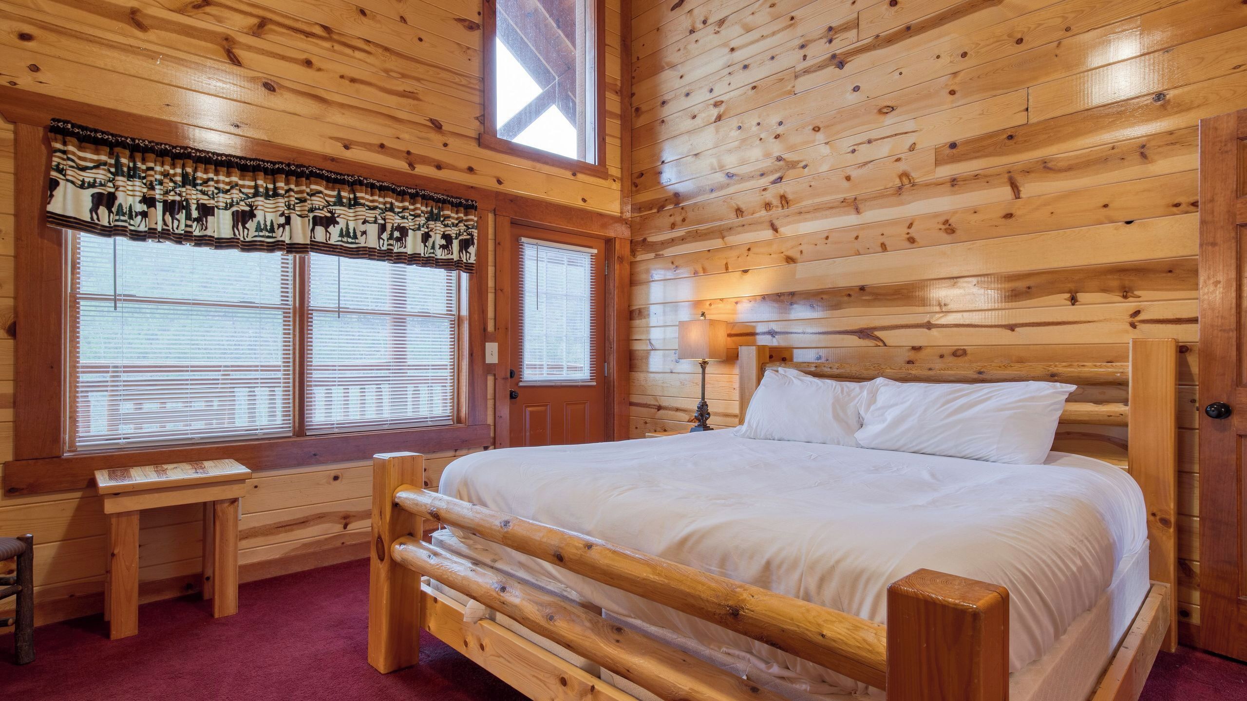 A wooden cabin bedroom with a bed, a nightstand, a lamp, and a window with blinds.