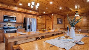 Une cuisine aménagée dans le style d’un chalet en bois, avec une longue table à manger dressée pour le repas, équipée d’appareils modernes et agrémentée d’un lustre.