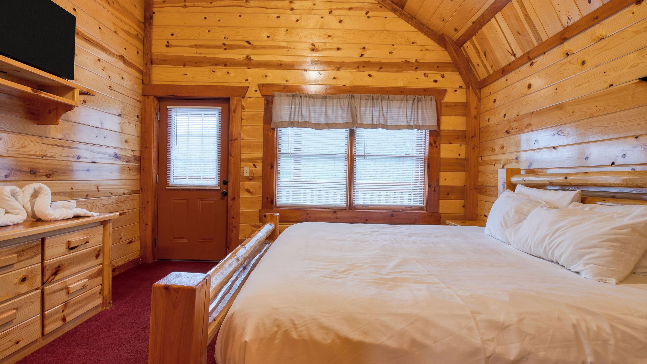 Una habitación de cabaña de madera con una cama, un tocador, una puerta y ventanas.