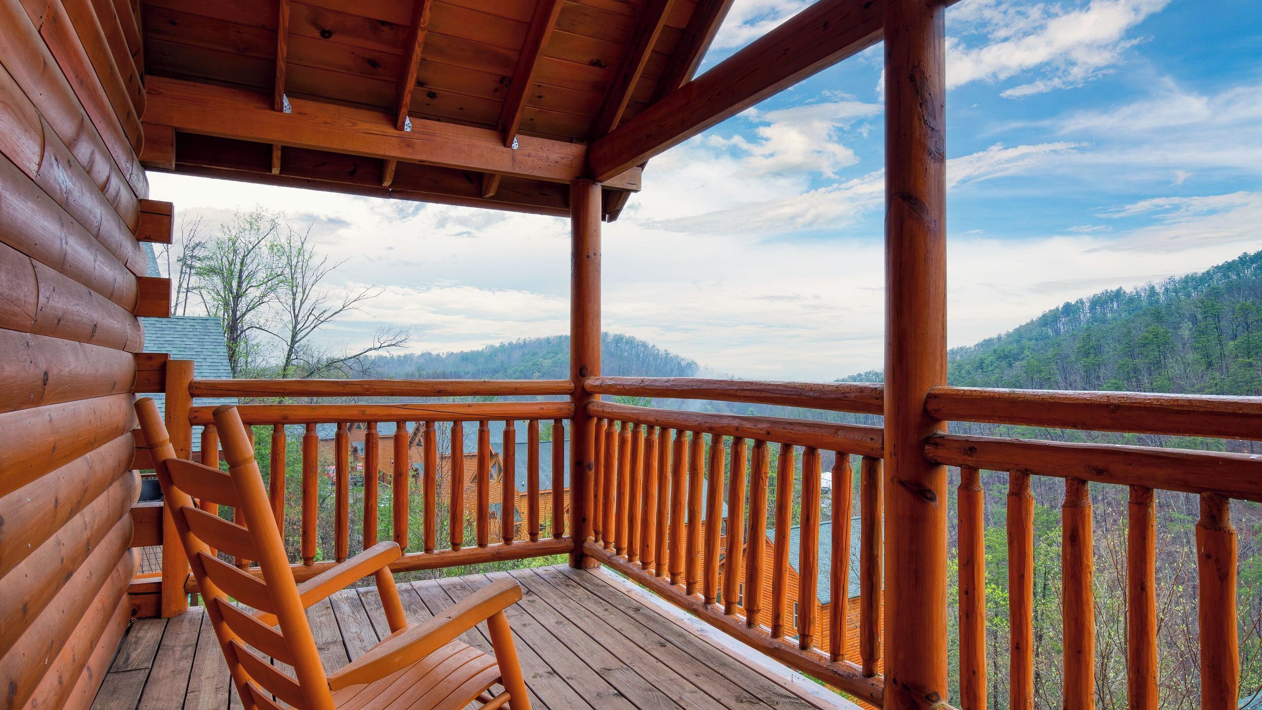 A wooden deck with a wooden chair, a wooden railing, and a view of a forested mountain range.