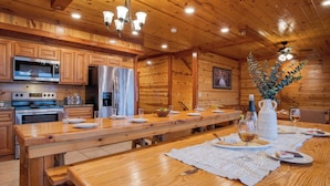 A wooden cabin-style kitchen with a long dining table set for a meal, equipped with modern appliances and a chandelier.