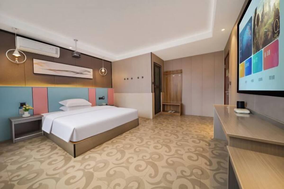 Una habitación de hotel moderna con cama, mesita de noche y una pantalla de televisión mostrando contenido colorido.